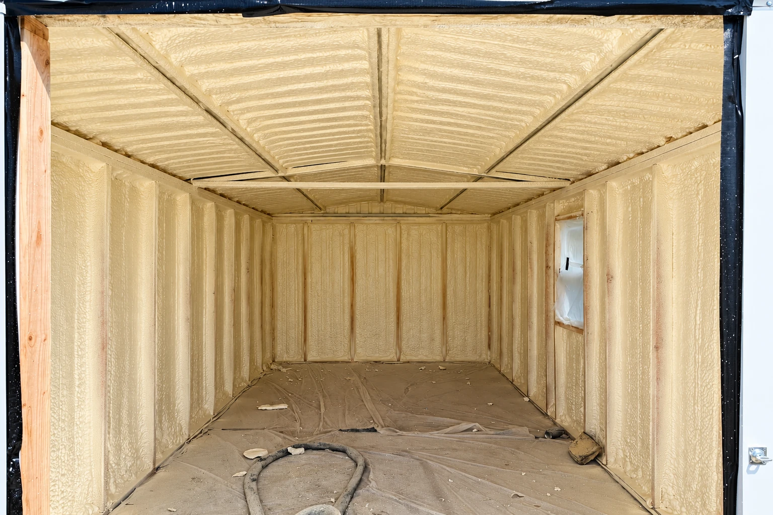 A small framed structure is coated inside with tan spray foam across its walls and pitched ceiling, with protective sheeting on the floor.